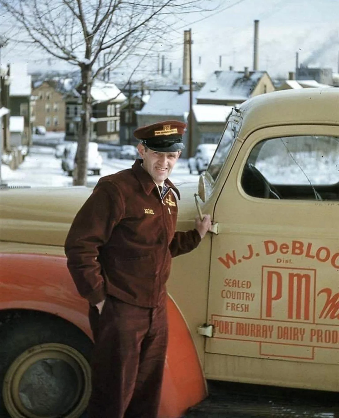 A uniformed milkman stands by a vintage Port Murray Dairy Products truck on a snowy street with houses and trees in the background.
