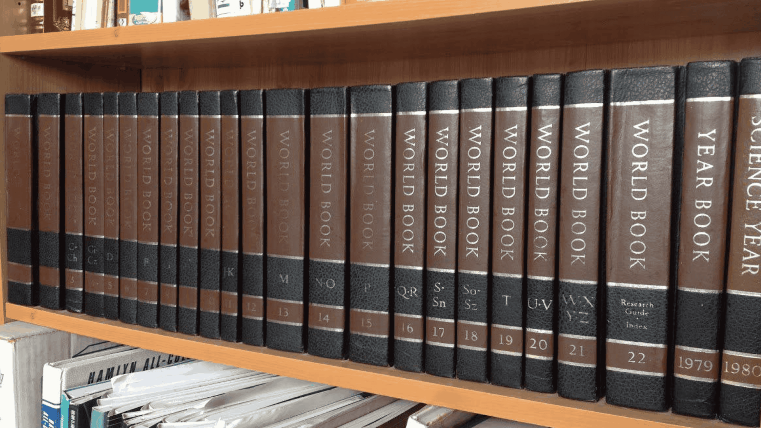 A row of World Book encyclopedias with brown and black spines is neatly arranged on a wooden shelf, alongside a few other books and binders.