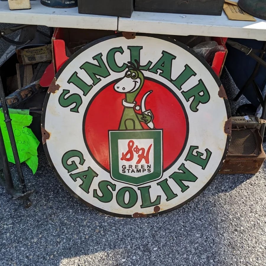 A round vintage Sinclair Gasoline sign features a cartoon dinosaur holding S&H Green Stamps, with a red background and green text, lying on pavement at a flea market.