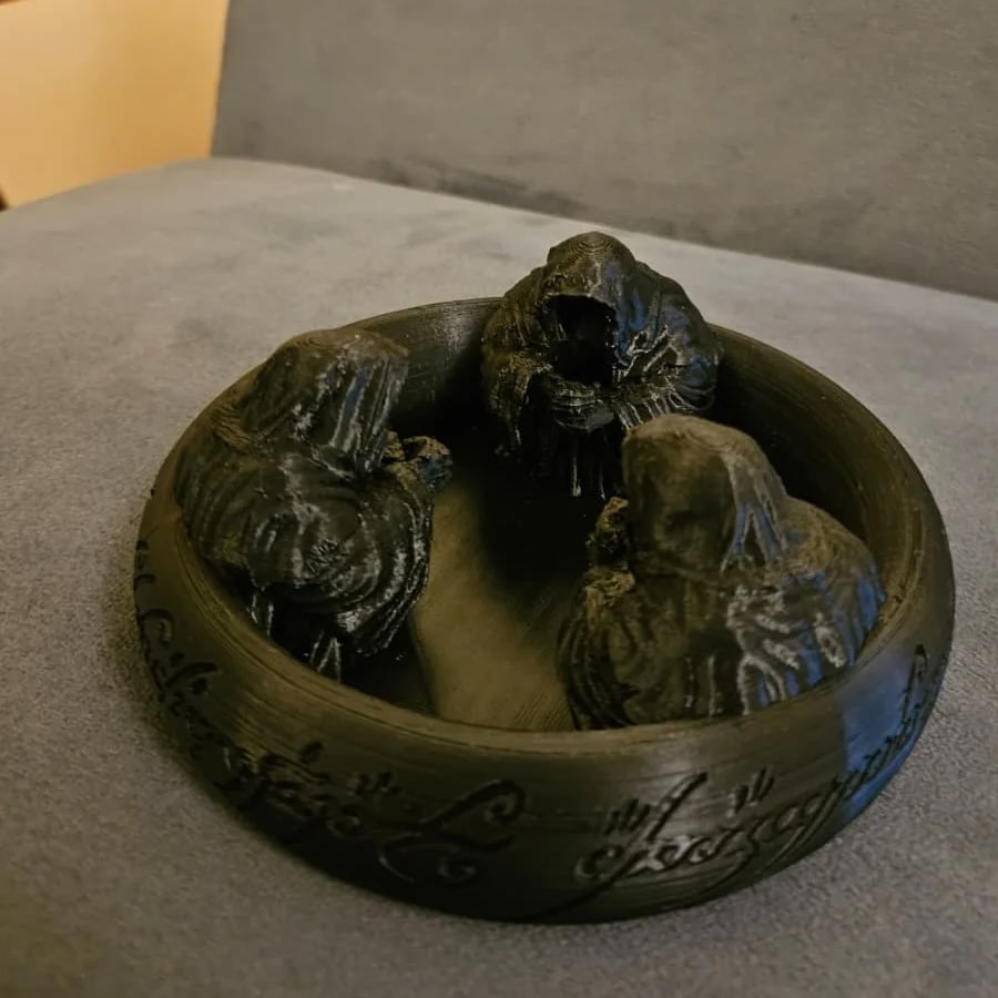 A black bowl-like object featuring three hooded, cloaked figures seated inside, with Elvish-style script engraved around the outer rim.