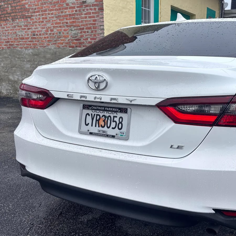 A white Toyota Camry LE parked on wet pavement near a brick and stone wall, displaying a Georgia license plate reading “CYR3058.”