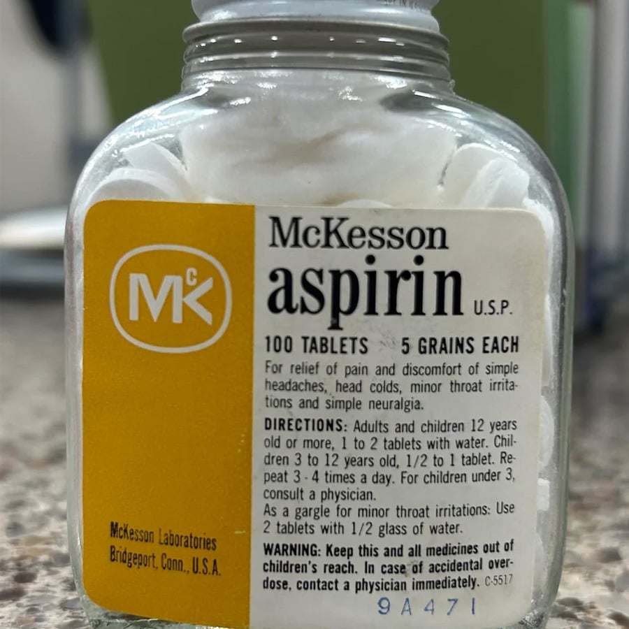 A close-up of a vintage McKesson aspirin bottle with white tablets inside, dosage instructions, and warnings printed on the yellow and white label.