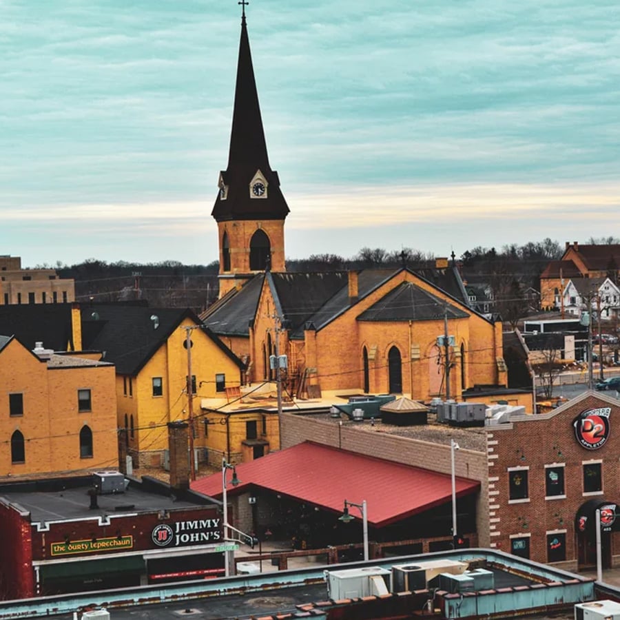 A cityscape featuring a large yellow brick church with a tall steeple, surrounded by various buildings, including a Jimmy John’s and a Pizza restaurant under a cloudy sky.