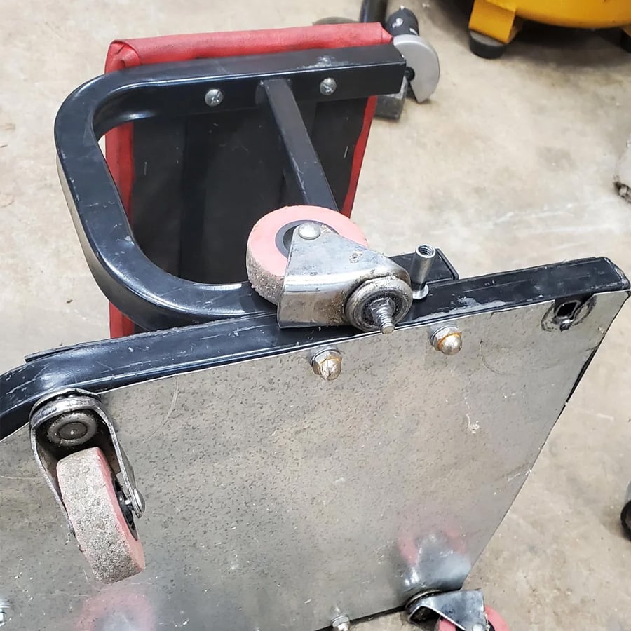 A close-up of a metal creeper seat with red and gray caster wheels, showing one detached wheel resting on its frame, on a concrete floor.