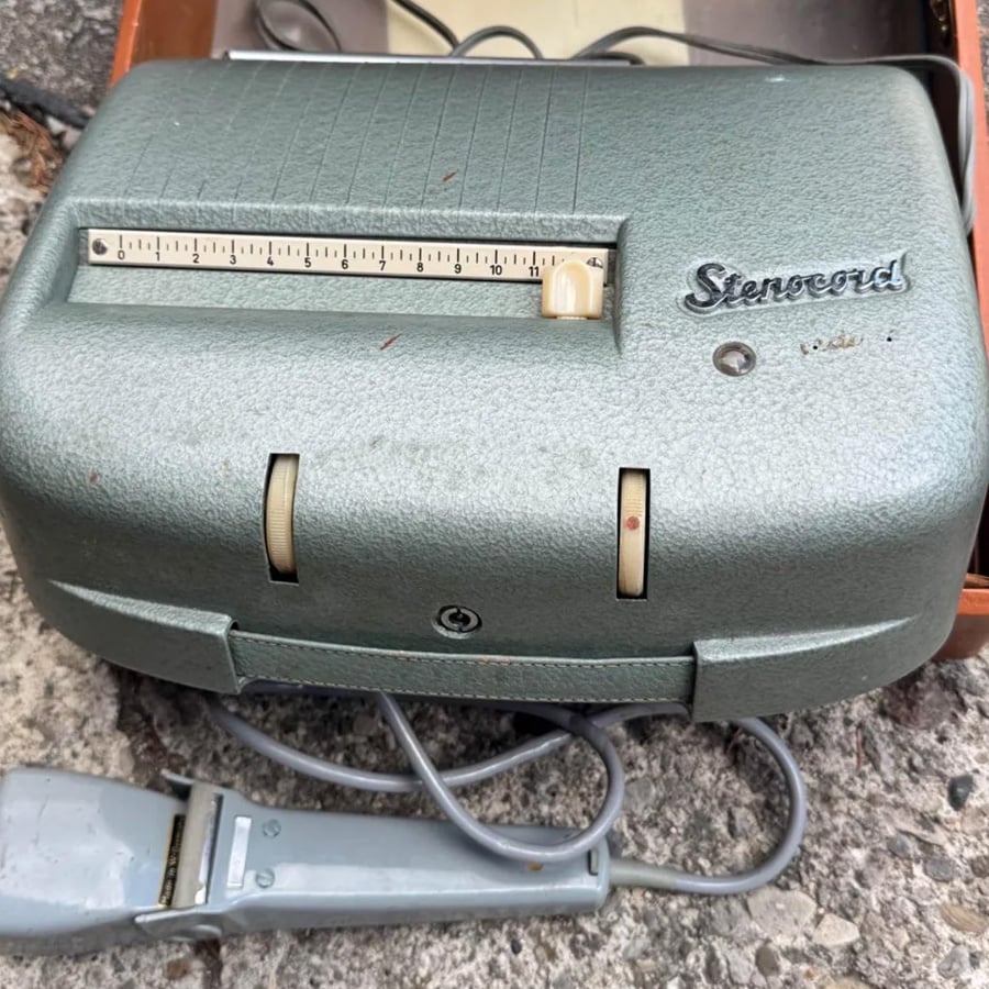 A vintage Stenograph machine with a ruler and sliders on top, the word “Stenocard” labeled, and an attached gray corded device, resting on a rough stone surface.
