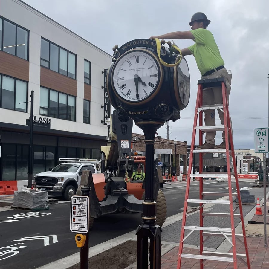 A worker on a red ladder cleans or repairs a large black street clock on a city sidewalk; construction vehicles and orange barriers are visible in the background.