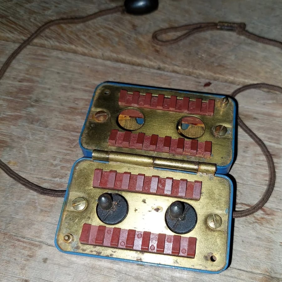 A vintage metal and plastic device with red buttons, two dials, and cords, laying open on a wooden surface. It appears to be an old electrical or communication tool.