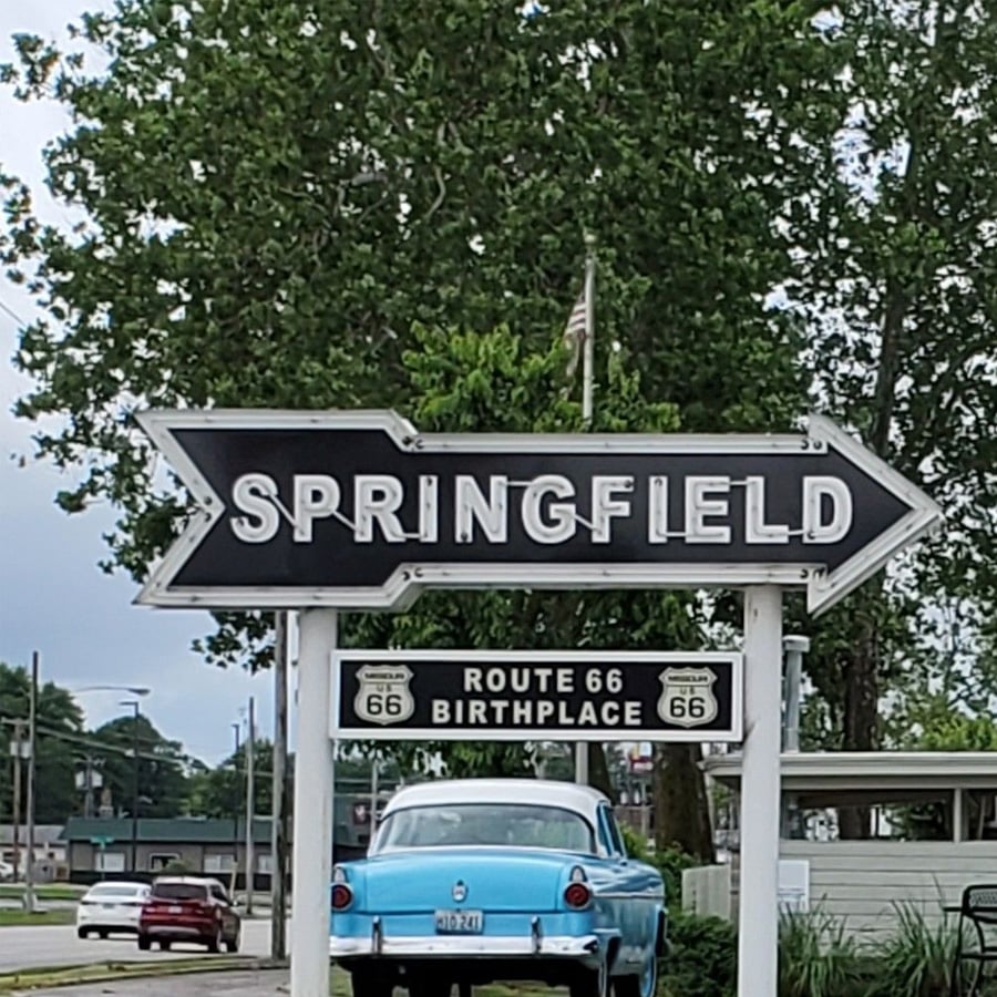 Large black-and-white signs point to Springfield and indicate it’s the birthplace of Route 66, with a vintage blue car parked underneath and trees, buildings, and a cloudy sky in the background.