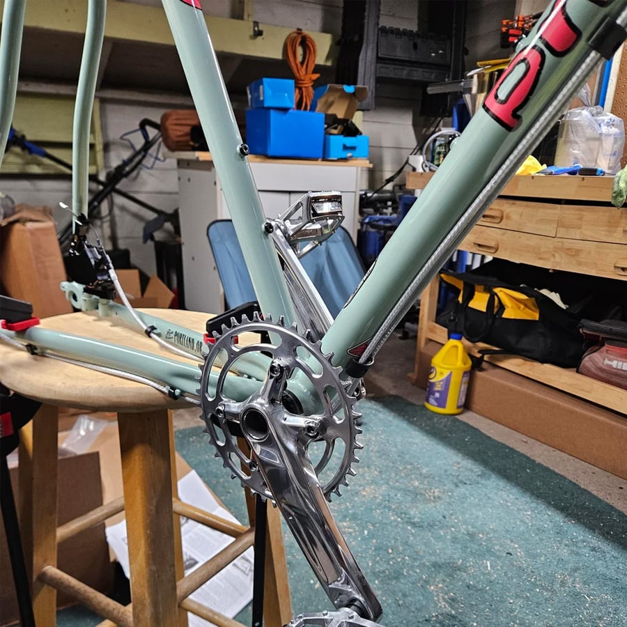 A partially assembled bicycle frame on a stool in a cluttered workshop, showing a crankset and front derailleur installed. Tools, boxes, and supplies are visible in the background.