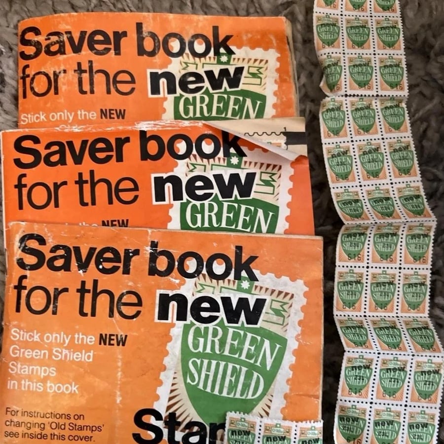 Three vintage “Green Shield” Saver Books are shown, each partially filled with Green Shield stamps. A strip of loose stamps is placed to the right of the books on a textured surface.