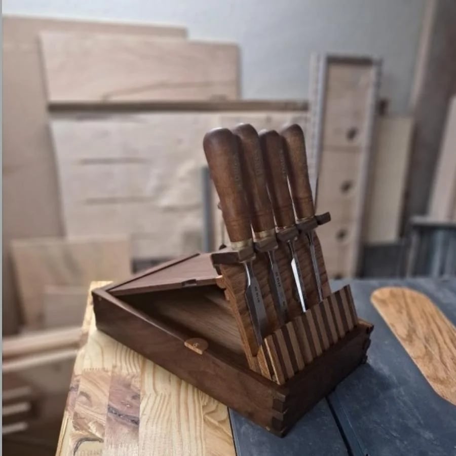 A wooden knife holder with four woodworking chisels is on a table in a workshop, surrounded by pieces of wood and unfinished wooden boards in the background.