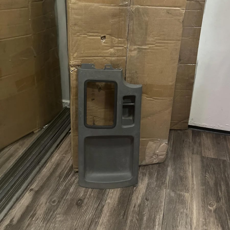 A gray plastic car door panel rests upright against a large cardboard box on a dark wood floor. The panel has a rectangular cutout and handle area.