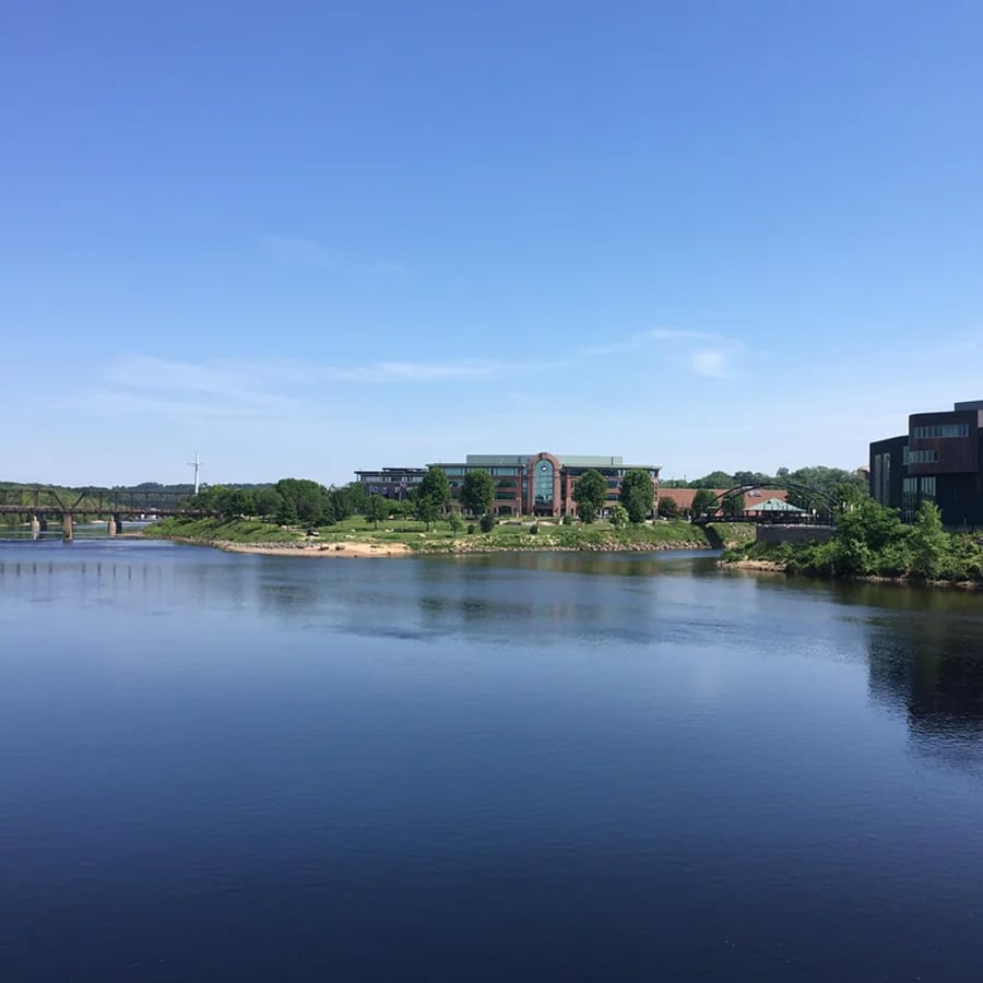 A calm river with a tree-lined bank, a few buildings in the center and right, a bridge to the left, and a clear blue sky above.