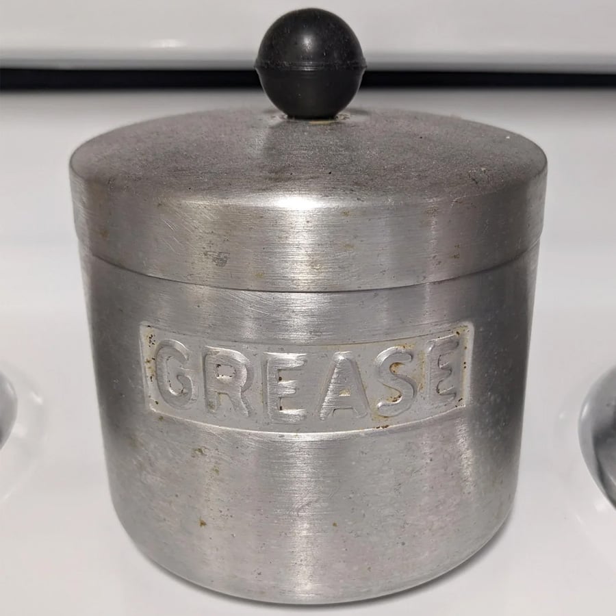 A round, metal container with a lid and black knob, labeled “GREASE” on its side. The container shows slight wear and sits on a white surface.