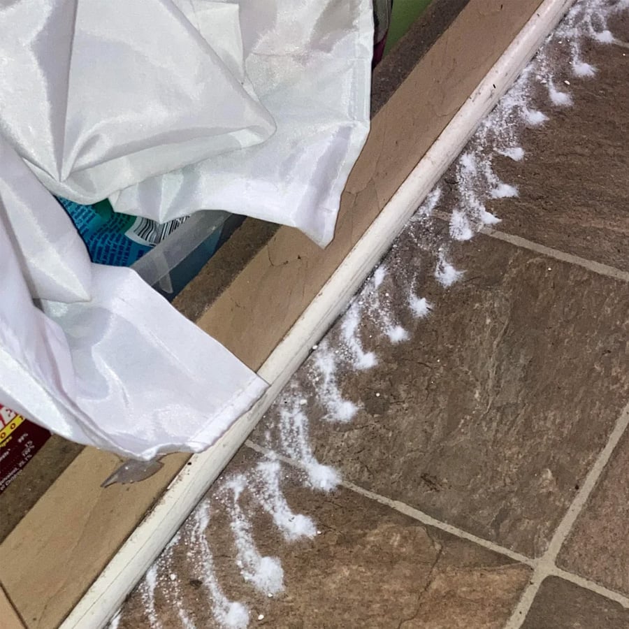 A line of white powder, possibly a cleaning agent or pest control, is spread along the edge of a tiled floor near a wall and under a partially draped white curtain.
