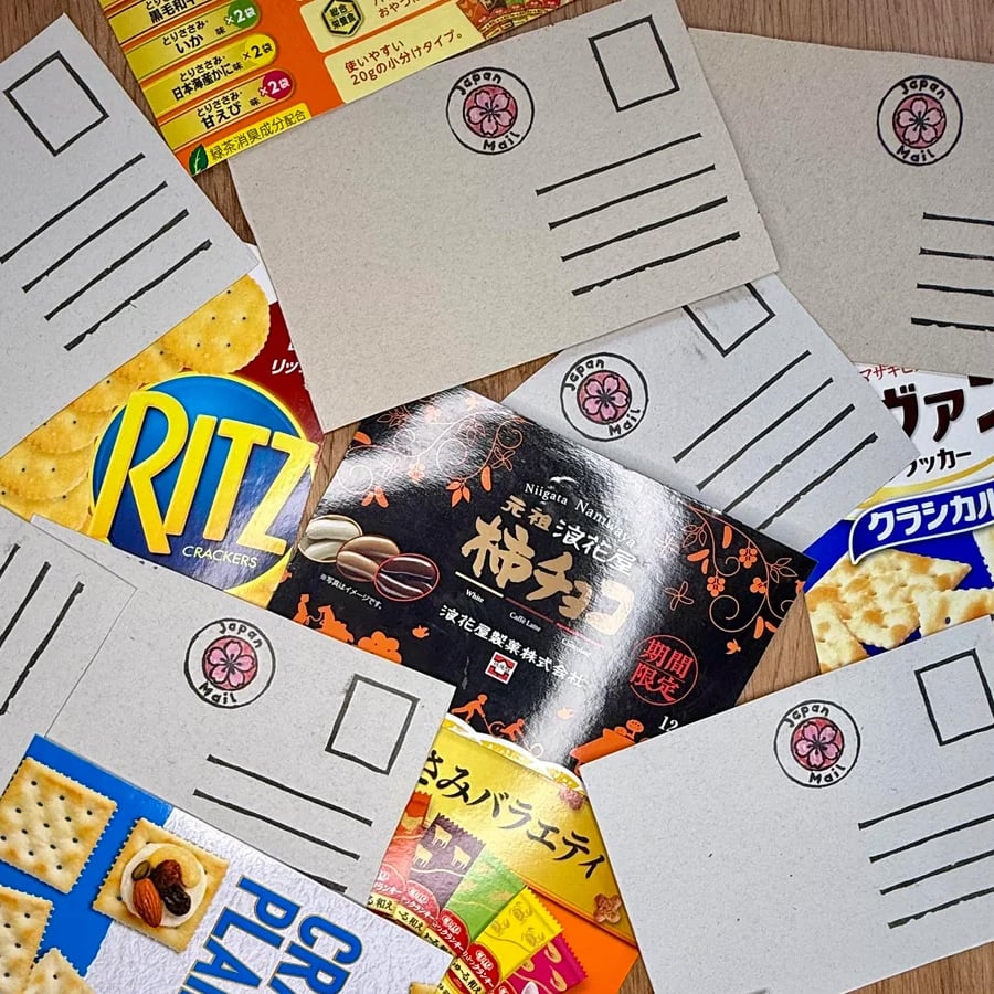 Several blank postcards with a cherry blossom emblem are placed on top of colorful Japanese snack packaging, including Ritz crackers and various other snacks, creating a lively and playful arrangement.