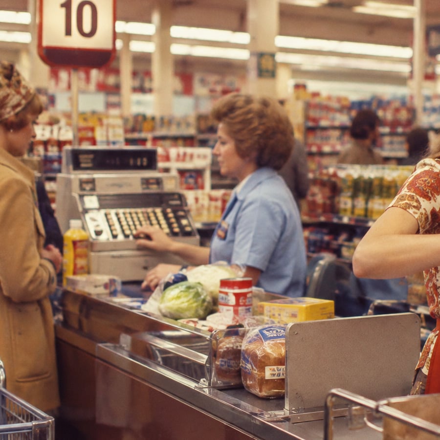 A cashier in a blue uniform rings up groceries for a customer at a supermarket checkout. A register displays the number 10 above. Various food items are on the conveyor belt.