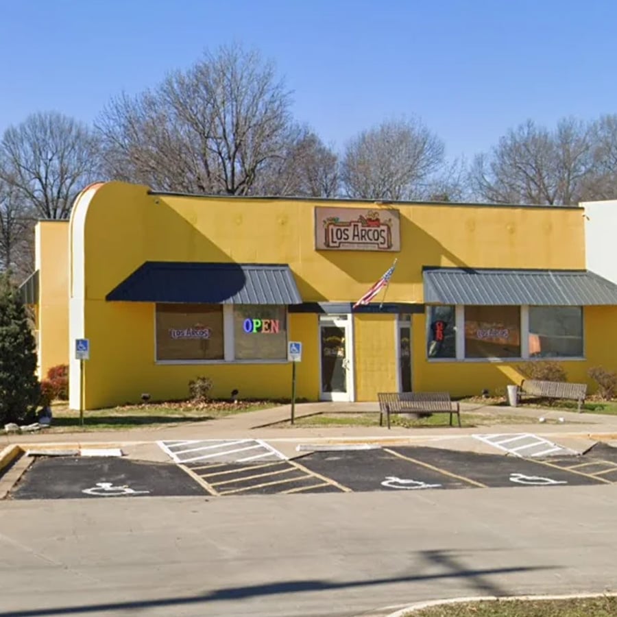 A yellow restaurant building with a sign reading “Los Arcos,” large windows, and an “OPEN” sign. There are benches out front, wheelchair parking spaces, and trees without leaves in the background.
