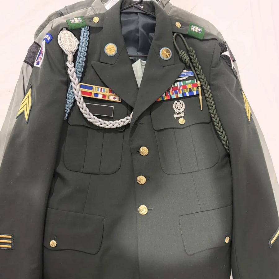 A decorated dark green military dress uniform with various ribbons, medals, patches, pins, and cords hanging on a hanger against a light background.