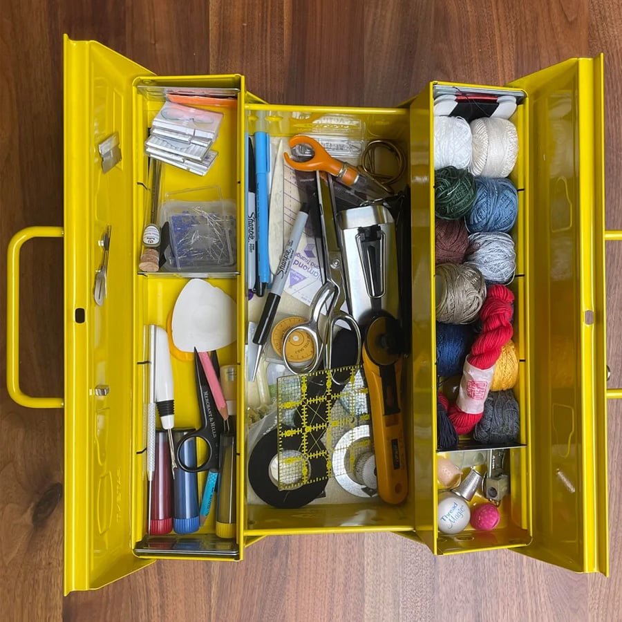 A yellow sewing box with open compartments displaying threads, scissors, marking tools, needles, pins, a measuring tape, a rotary cutter, and various sewing accessories organized neatly inside.