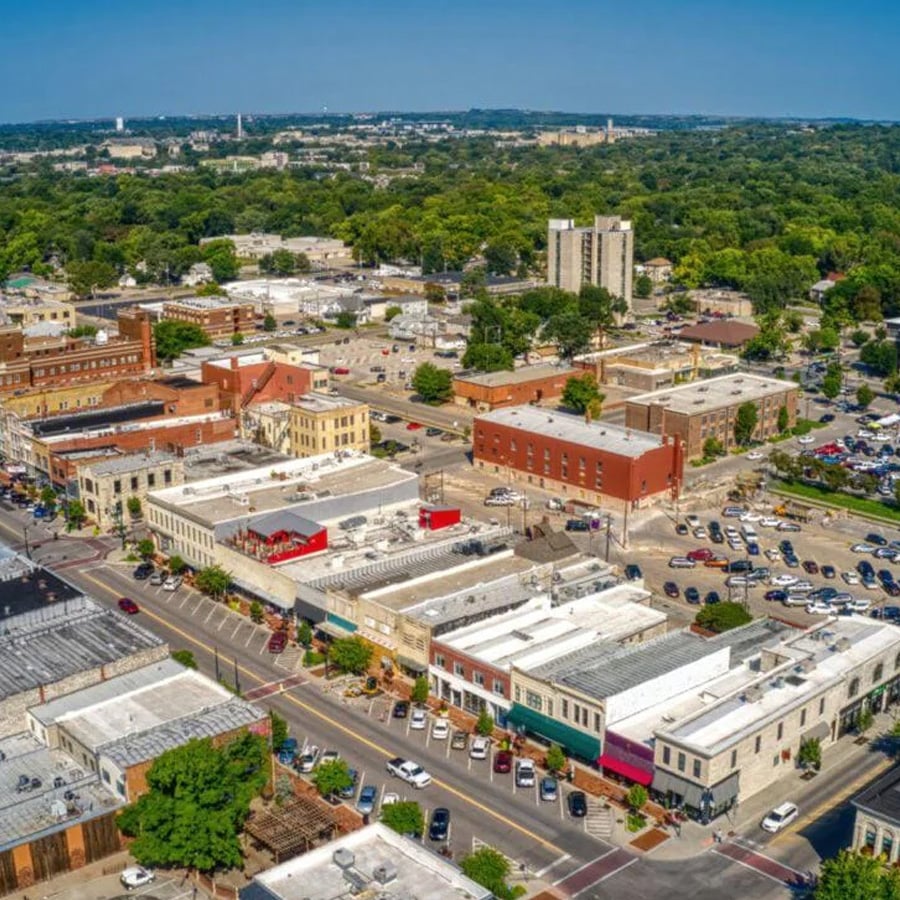 Aerial view of a small city downtown area with historic brick buildings, parking lots, streets, and surrounding green trees under a clear blue sky.