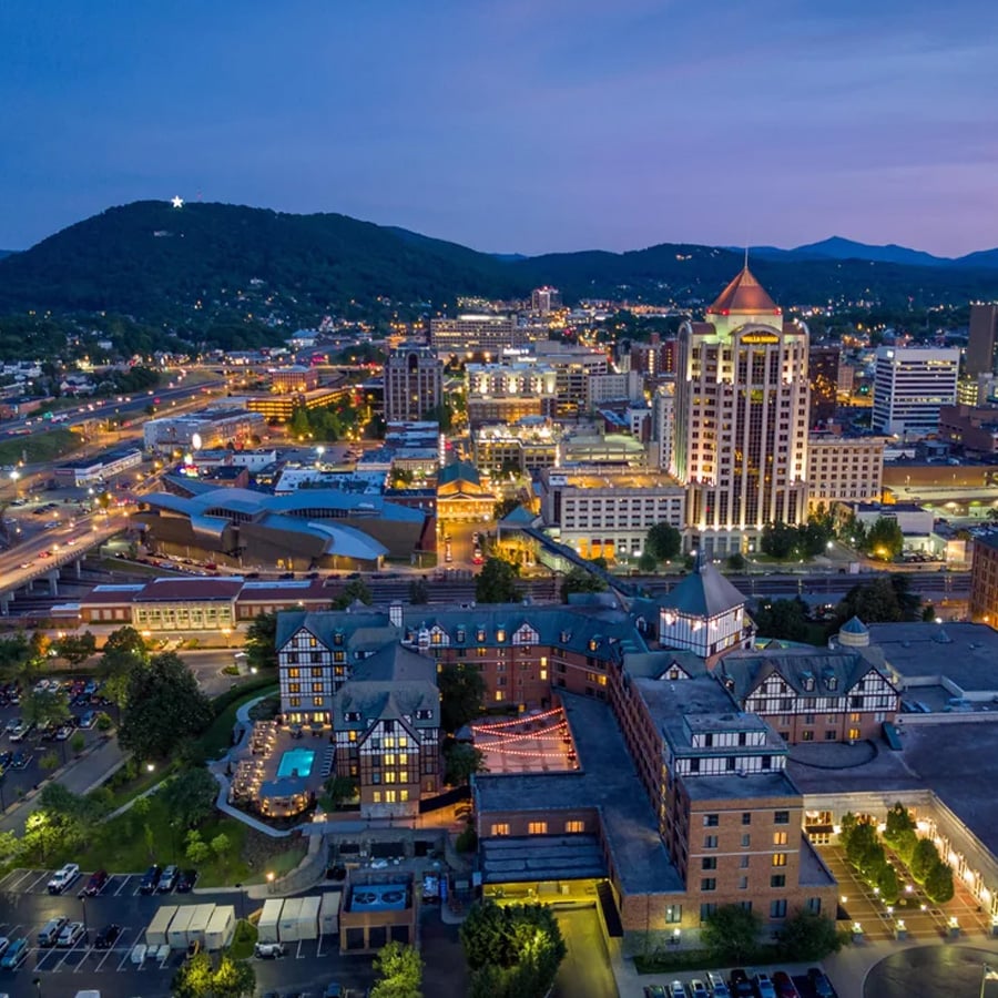 Aerial view of a city at dusk with illuminated buildings, streets, and cars; mountains and a lit hillside star are visible in the background. A hotel with a pool is seen in the foreground.