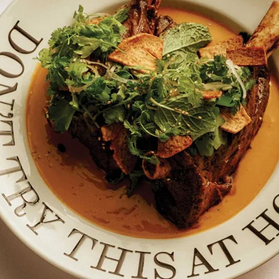 A plate with braised meat topped with fresh herbs and crispy chips in a golden sauce. The plate rim reads “DON’T TRY THIS AT HO.”