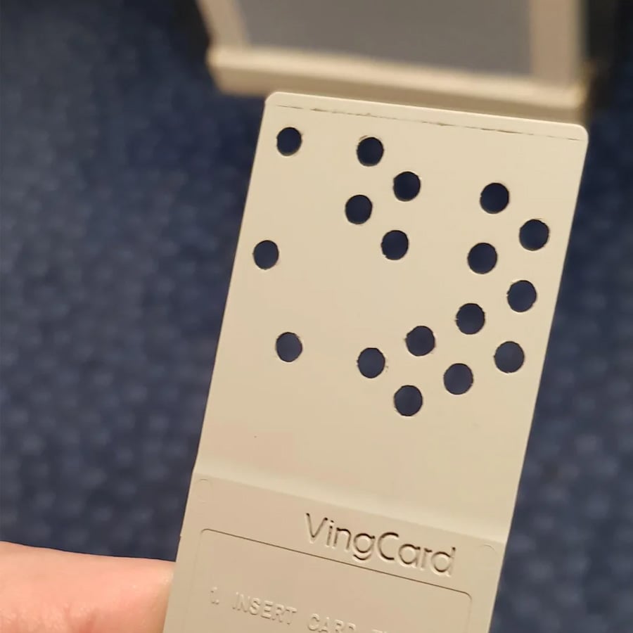 A hand holds a vintage VingCard hotel room key card with multiple round holes punched in a specific pattern. The background shows blue patterned carpet and part of a door.