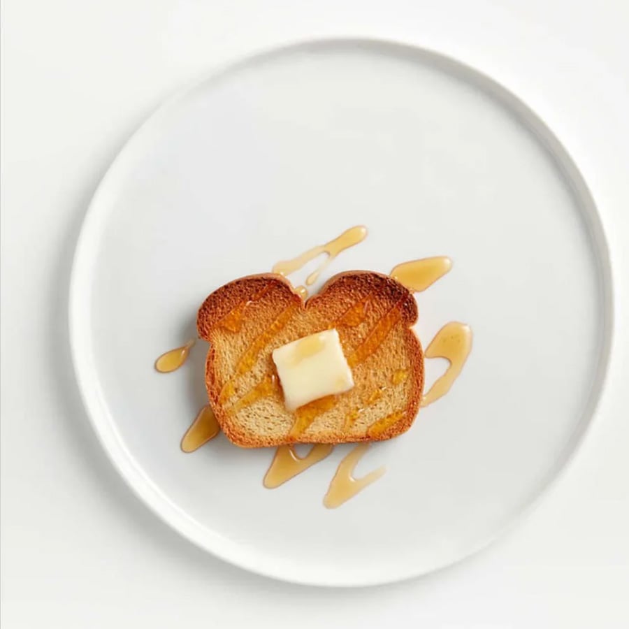 A slice of toasted bread with a pat of butter on top, drizzled with syrup, sits on a white plate against a white background.