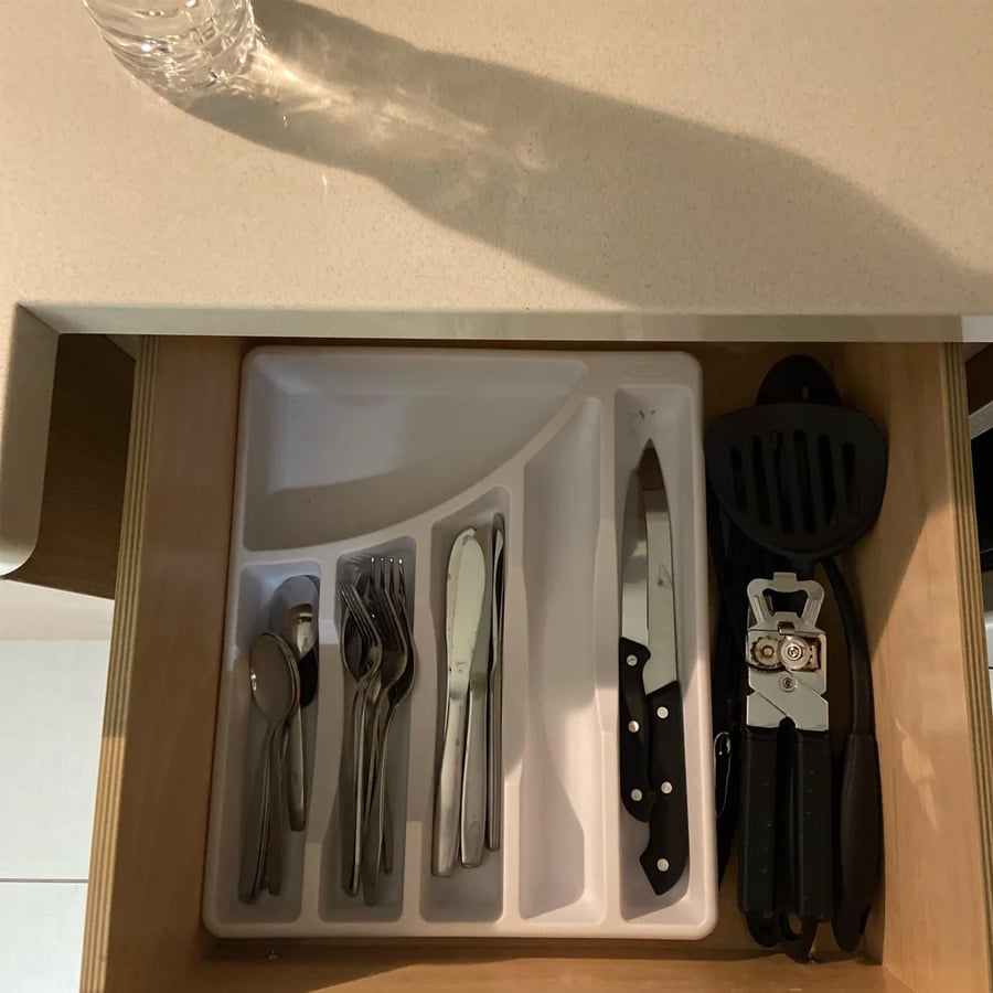 Open kitchen drawer with a white utensil organizer holding spoons, forks, knives, and a larger knife. Beside the organizer are a can opener and two black utensils. A water bottle is on the countertop above.