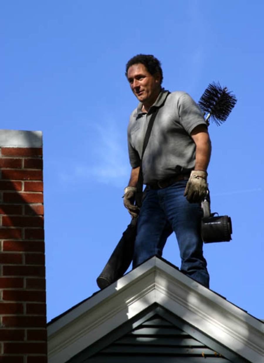 A man stands on a roof holding chimney cleaning equipment, including a chimney brush and a vacuum, next to a brick chimney, under a clear blue sky.