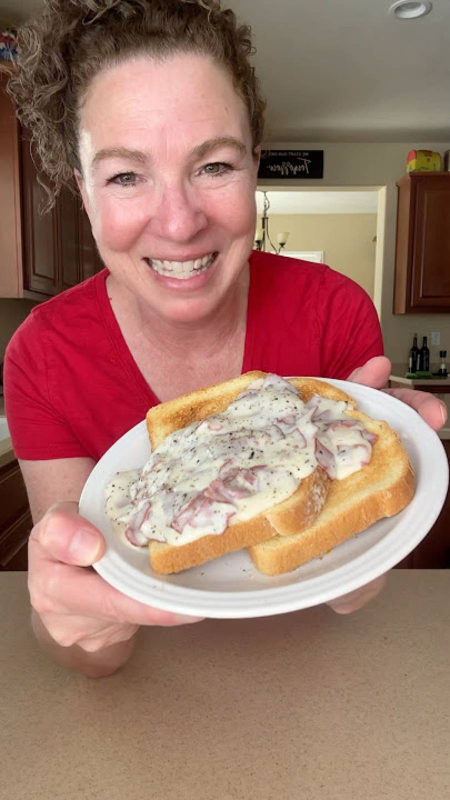 A smiling woman in a red shirt holds a plate with two slices of toast topped with a creamy, white gravy and pieces of meat, standing in a kitchen.