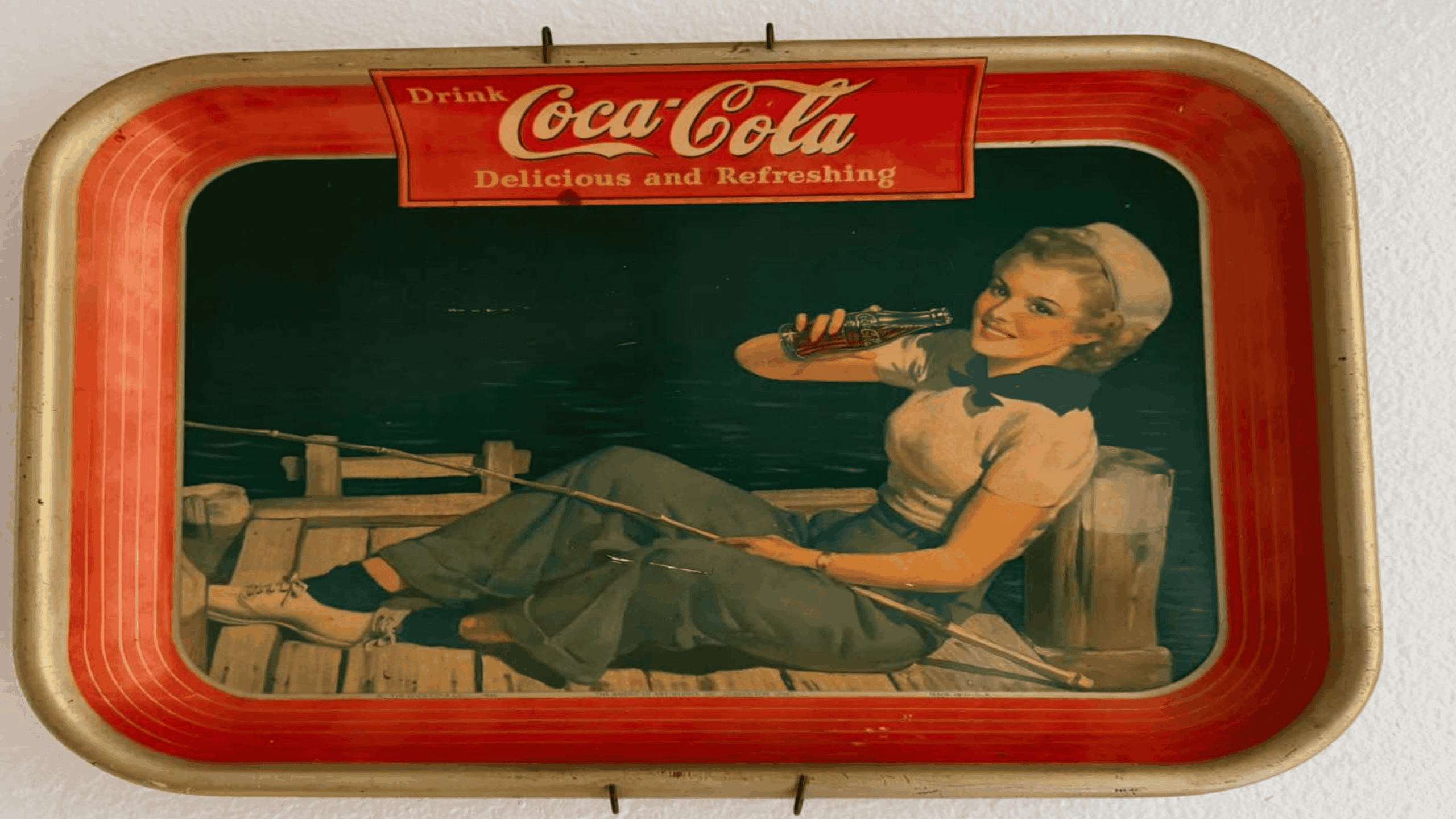 A vintage Coca-Cola tray shows a woman in sailor attire sitting on a dock, fishing and drinking a Coke. The tray is bordered in red and features the slogan “Drink Coca-Cola: Delicious and Refreshing” at the top.