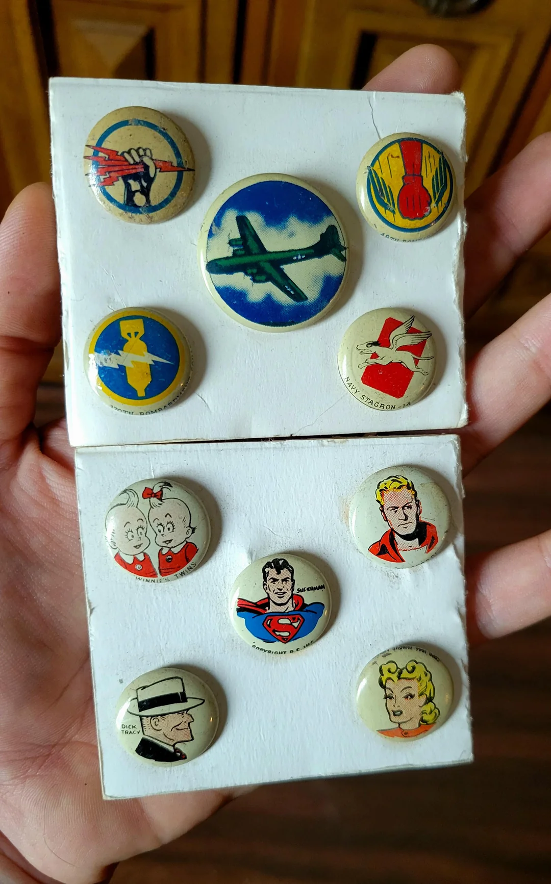 A hand holds two cards displaying vintage comic and cartoon pin-back buttons, featuring colorful drawings of various characters, symbols, and airplanes on a wooden background.