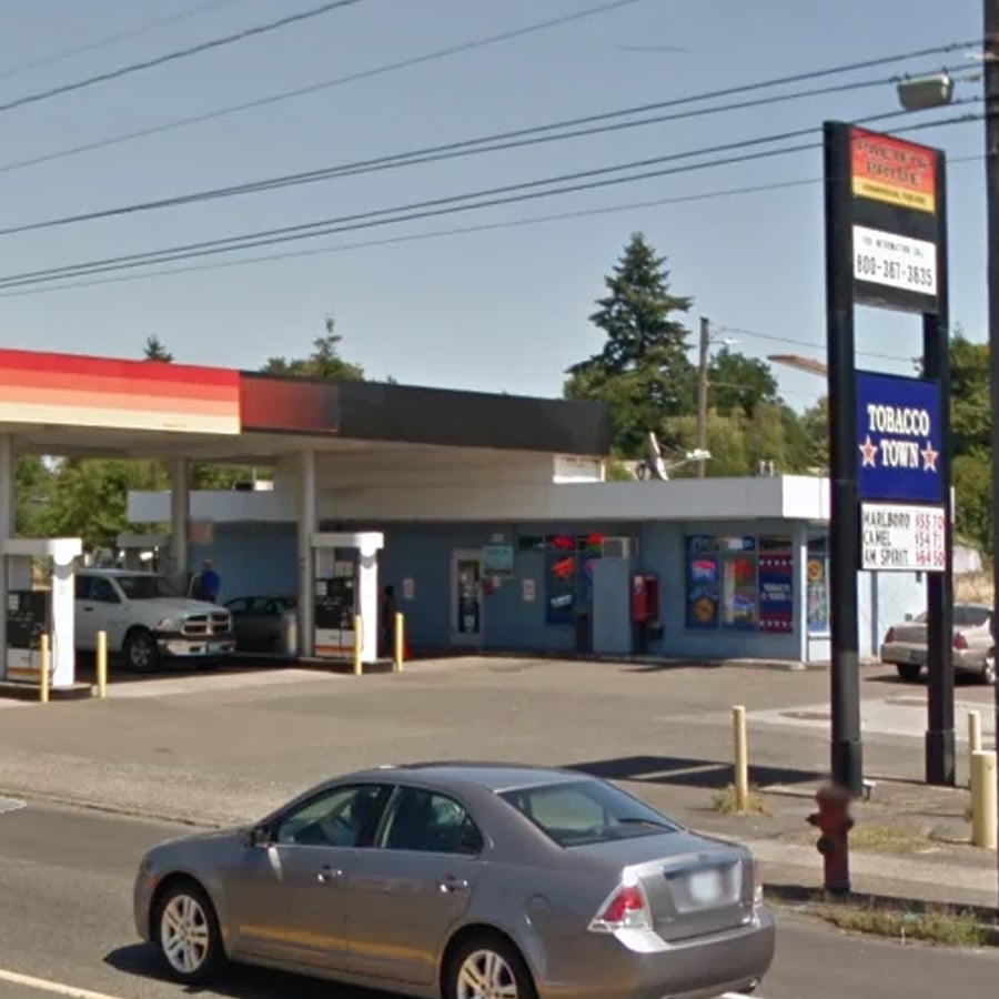 A silver car is parked in front of a small gas station and market. Several gas pumps are visible, and a tall sign reads “Tobacco Town” along with other information. Trees and power lines are in the background.