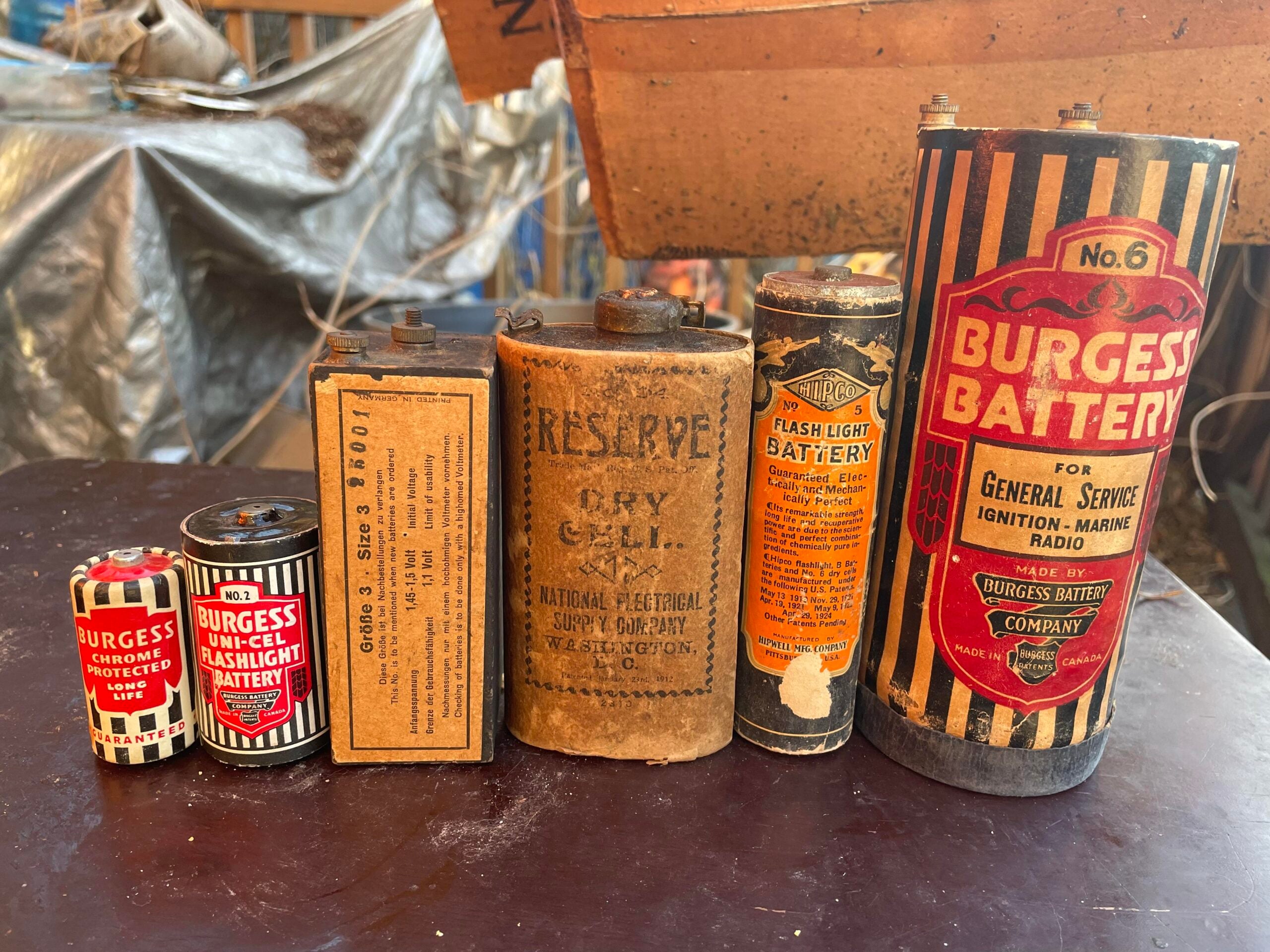 Six vintage batteries of varying sizes and labels, including Burgess brand and “Reserve Dry Cell,” are displayed upright on a dark surface in an industrial setting.