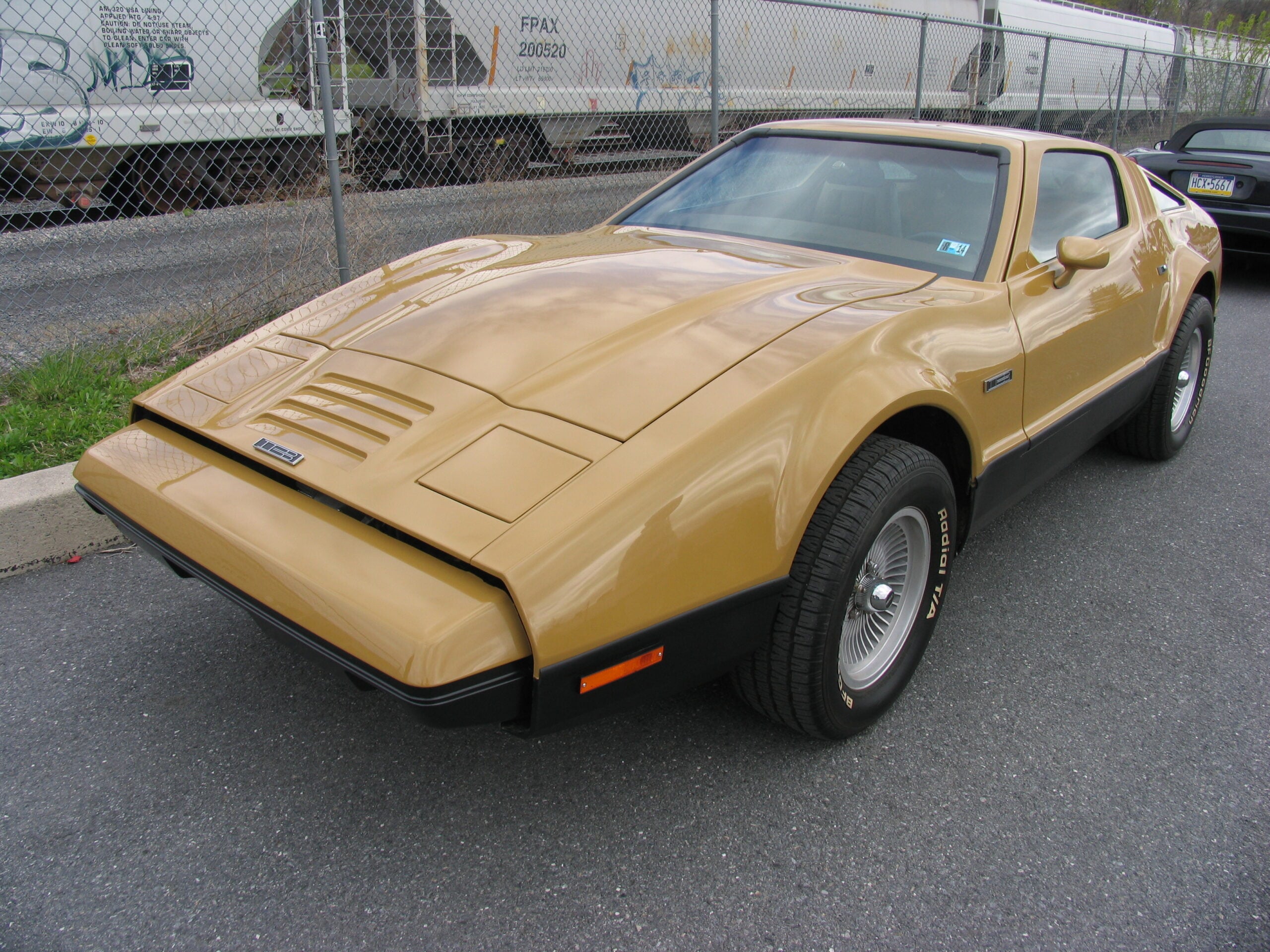 A gold-colored vintage car parked on a street beside a chain-link fence with a train in the background. The car has a sleek, angular design and black trim around the wheels.