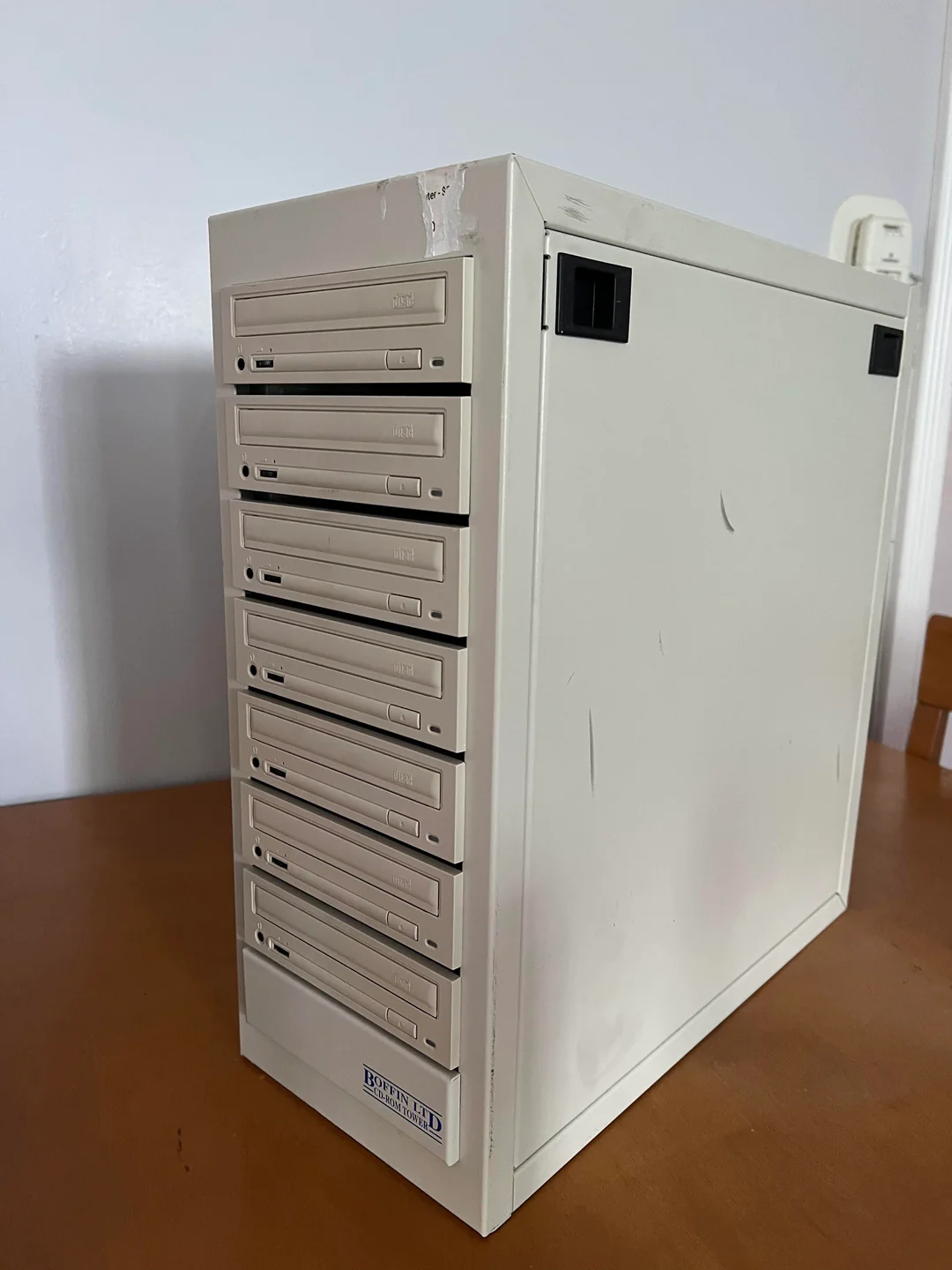 A beige computer tower with eight front-facing CD-ROM drives sits on a wooden table against a white wall. The case has some scratches and black vents near the top.