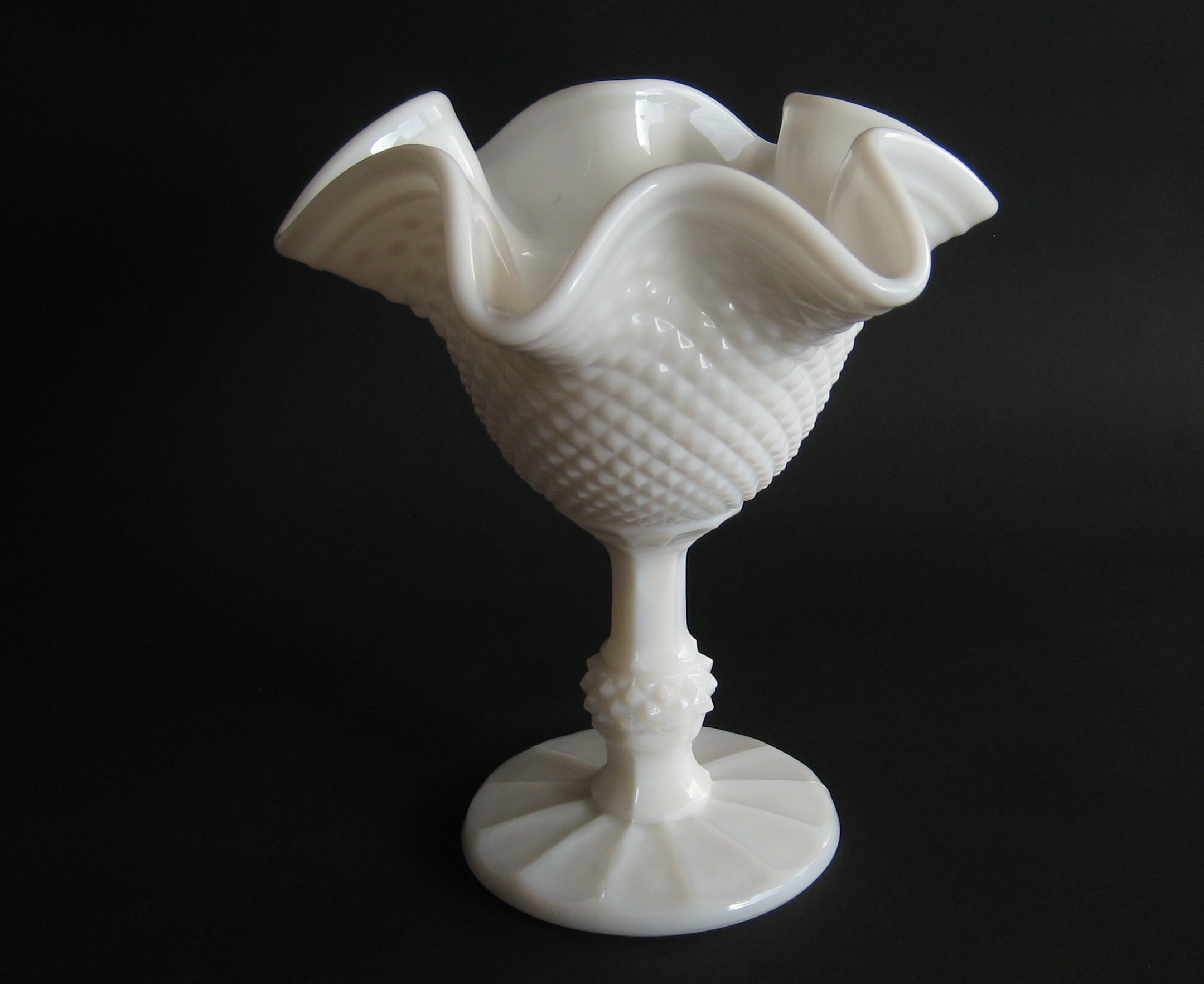 A vintage white milk glass vase with a ruffled edge and textured pattern, standing on a pedestal base against a black background.