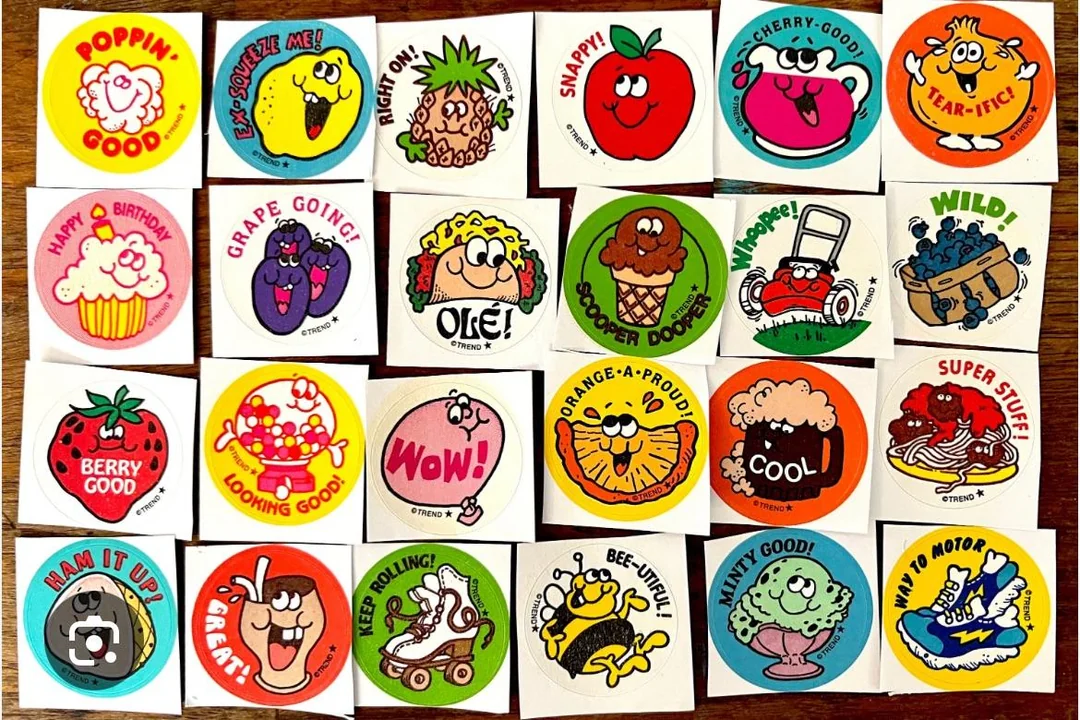 A collection of colorful, vintage reward stickers featuring cartoon fruits, desserts, and mascots with positive phrases like “Poppin’ Good,” “Berry Good,” “Bee-utiful,” and “Snappy!” arranged in a grid on a wooden surface.