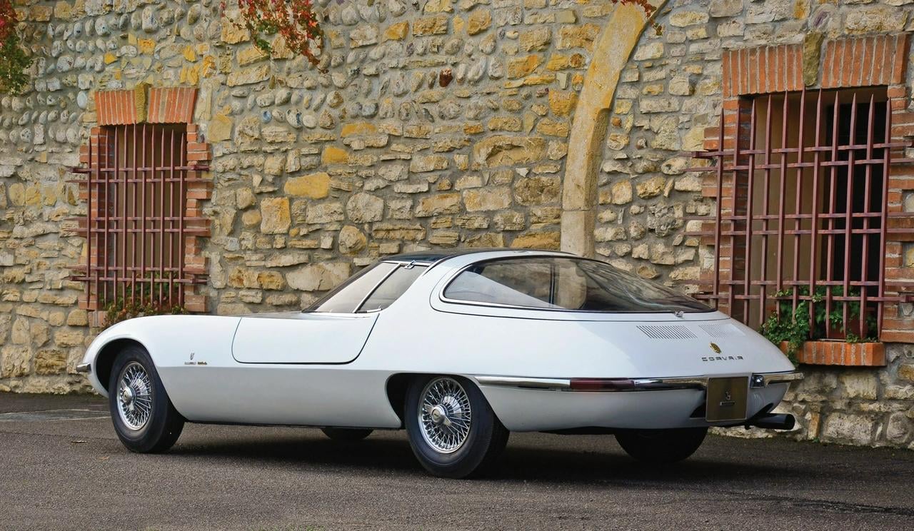 A vintage white sports car with a sleek, curved design is parked in front of a rustic stone wall with two barred windows framed by red bricks.