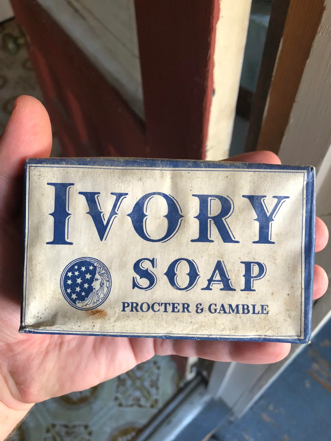 A hand holds a vintage bar of Ivory Soap by Procter & Gamble, featuring blue text and a logo with stars. The packaging appears old and slightly worn.