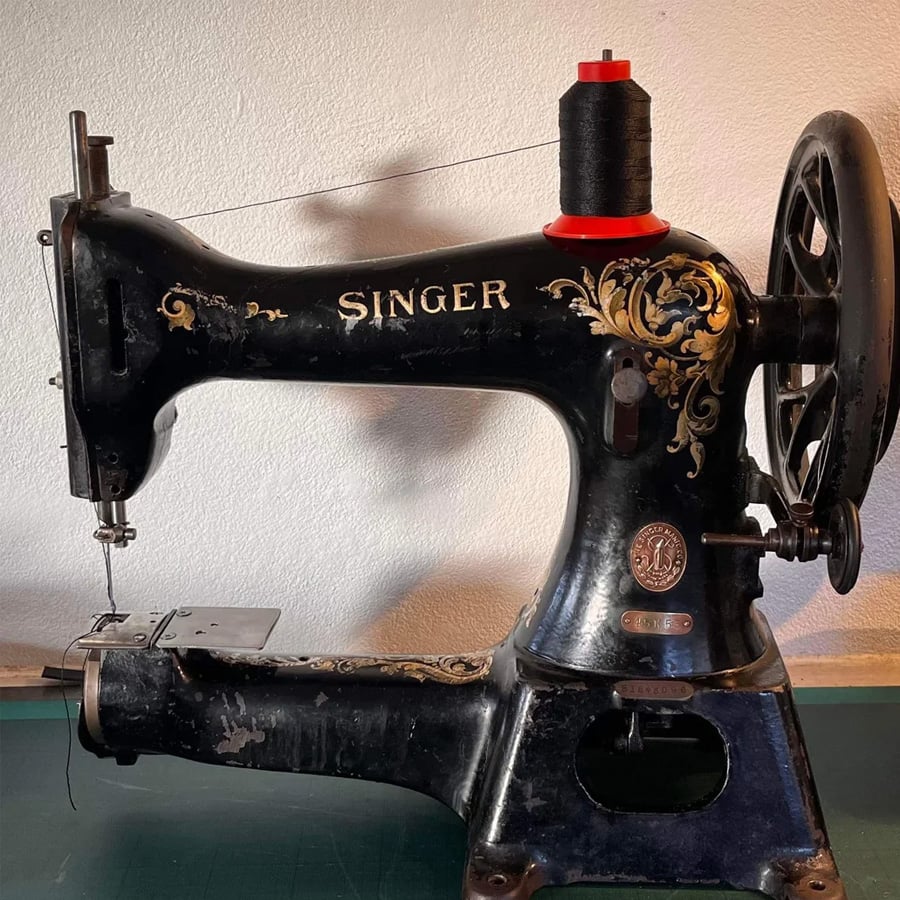 An antique black Singer sewing machine with ornate gold detailing, a spool of black thread on top, and a metal wheel on the right side, set against a plain wall.