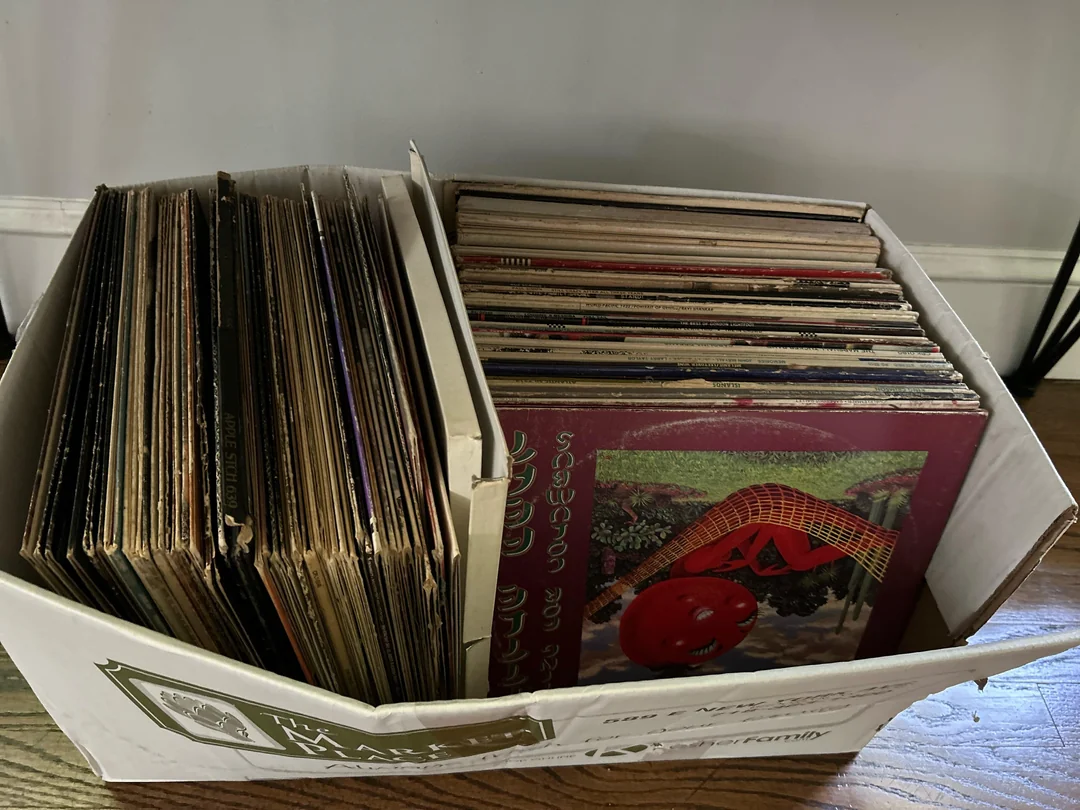 A white box filled with a large collection of vinyl records, some leaning to the left and others stacked vertically on the right, resting on a wooden floor against a white wall.
