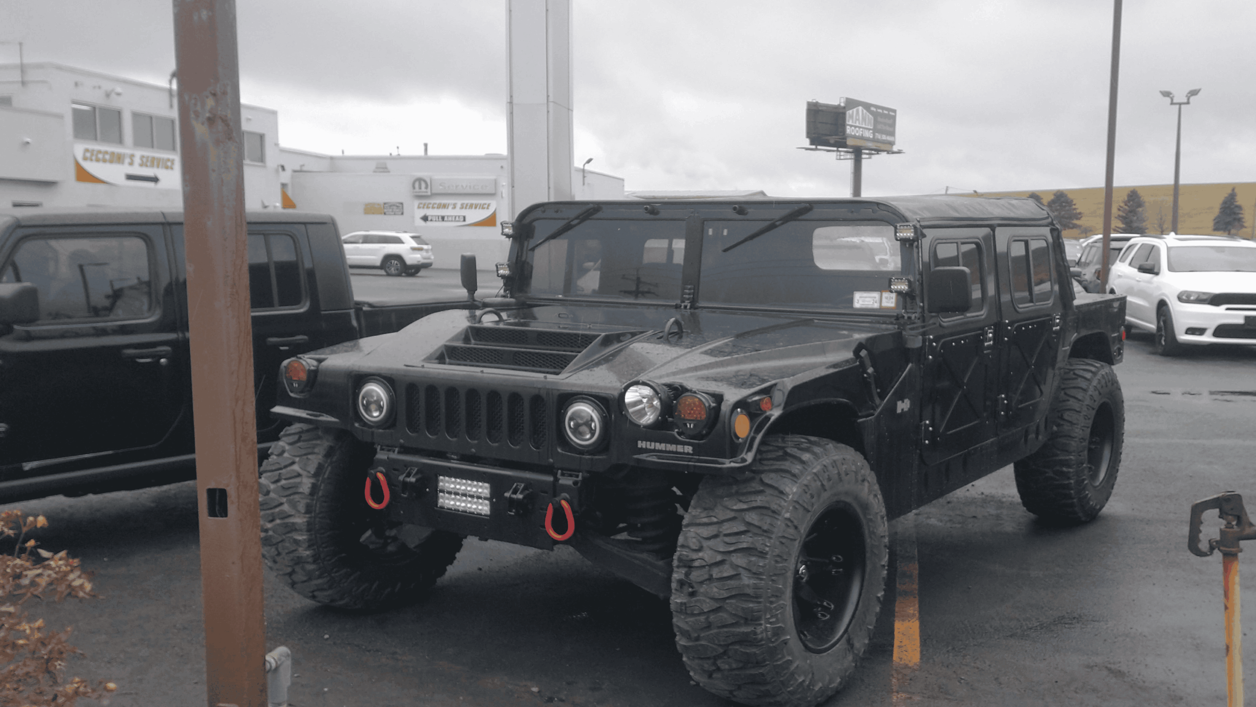 A black Hummer H1 with large off-road tires is parked in a lot on a cloudy day. Other vehicles and buildings are visible in the background.