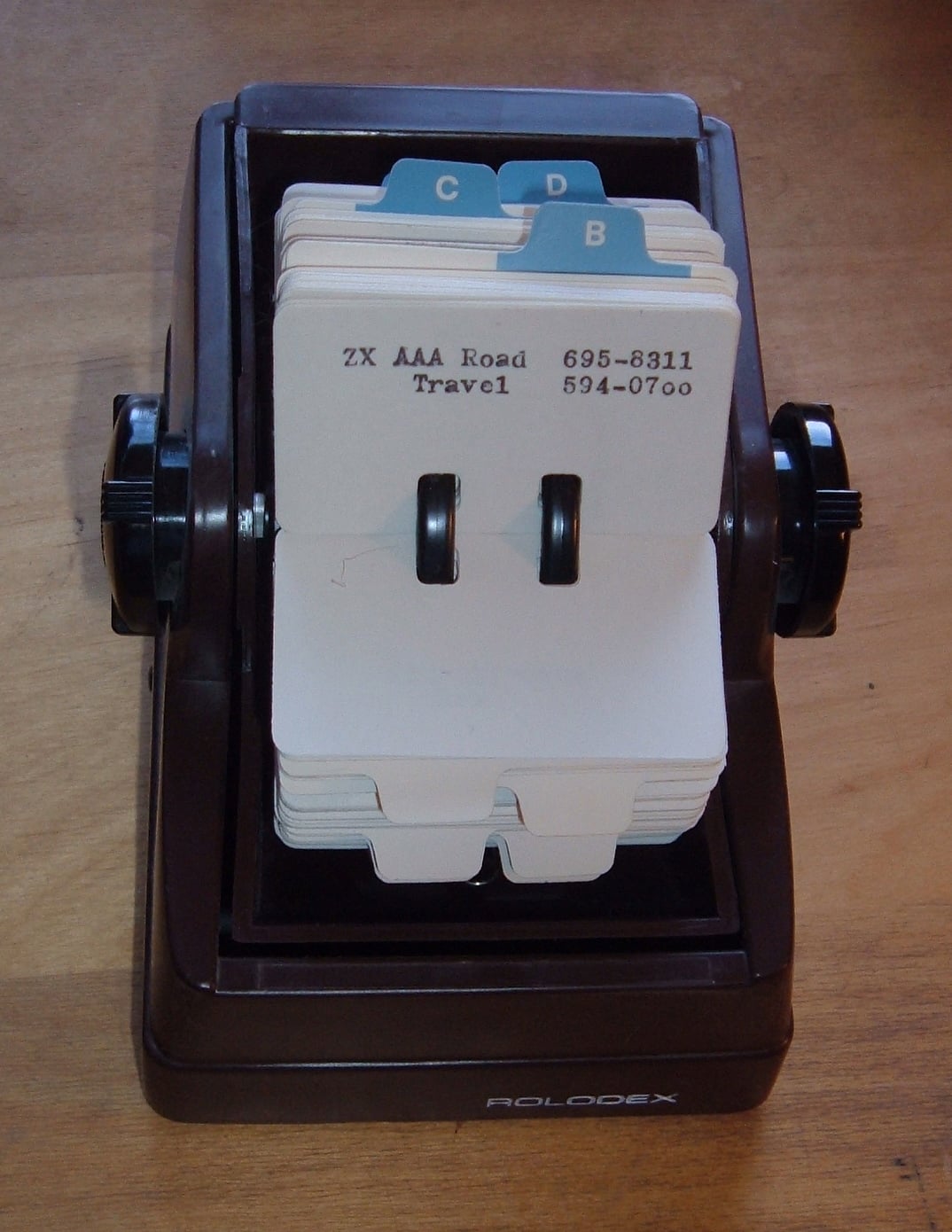 A Rolodex with alphabetical tabbed cards sits on a wooden surface. The cards display names and phone numbers, and the Rolodex is turned to cards labeled “B” and “C.”