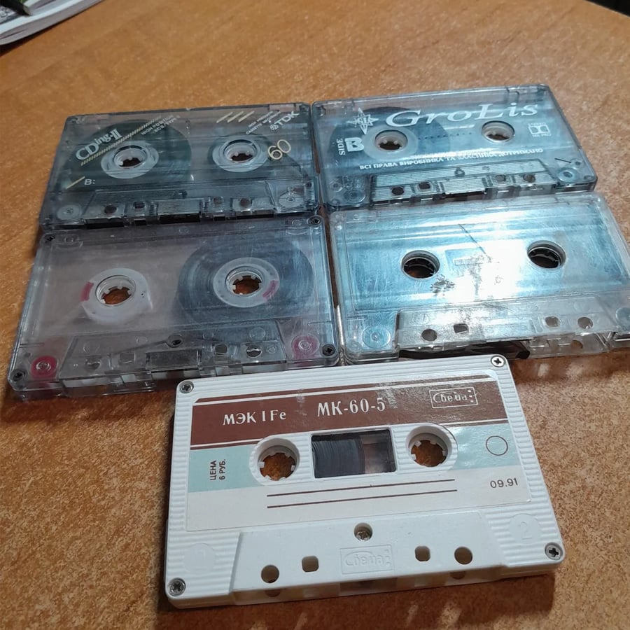 Five audio cassette tapes, four clear and one white, are arranged on a brown surface. The tapes have visible reels and labels with various brand names and markings.