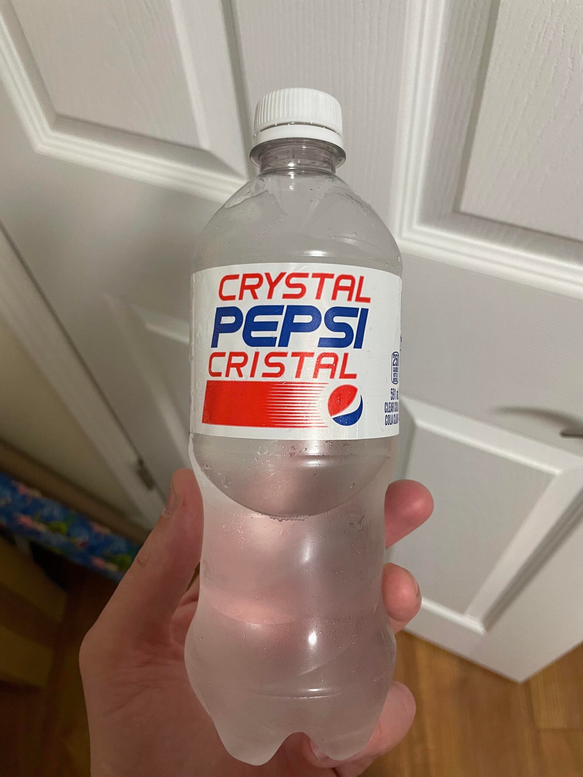 A hand holds a clear plastic bottle labeled “Crystal Pepsi Cristal” in front of white closet doors. The bottle contains a clear, colorless soda.