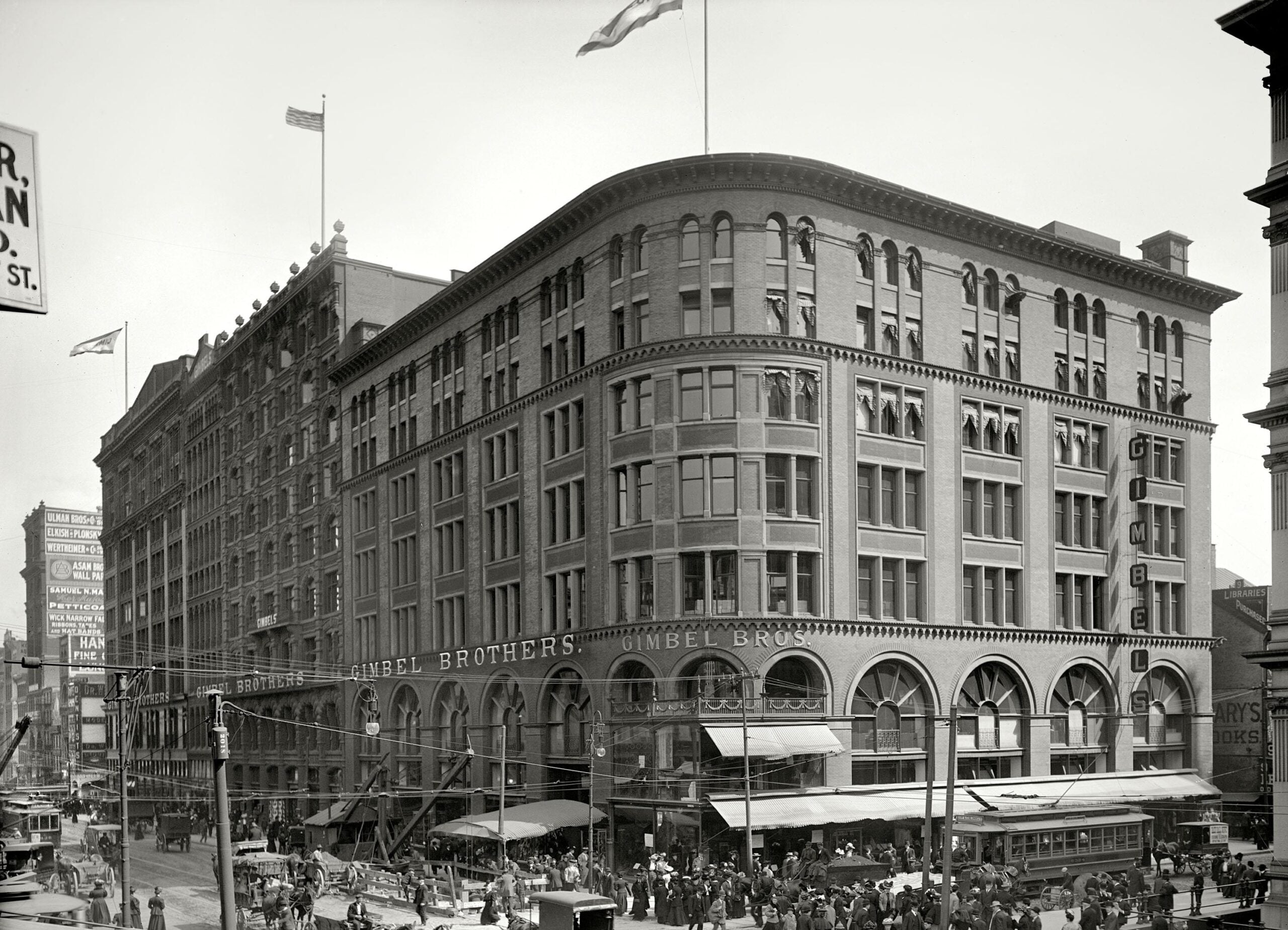 Historic black-and-white photo of a busy city street with crowds near a large multi-story brick building labeled “Gimbel Brothers,” featuring arched windows and multiple flags on the roof.