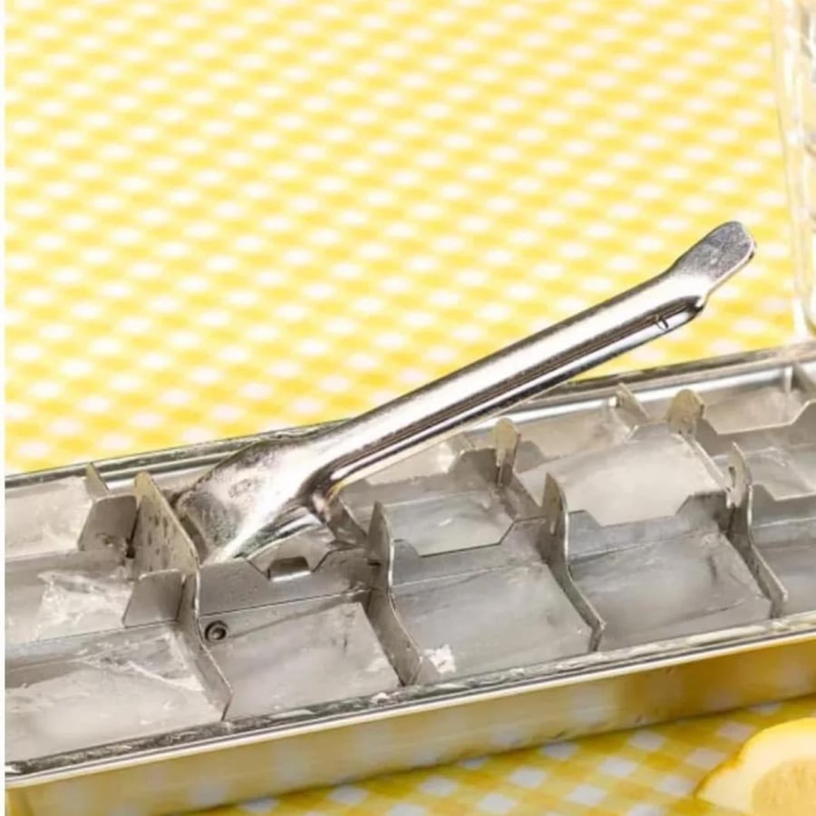 A vintage metal ice cube tray with a lever sits on a yellow and white checkered surface, partially filled with ice.